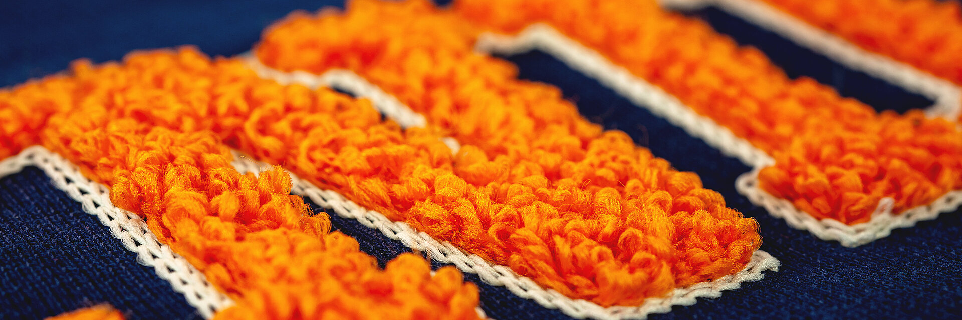 Nahaufnahme einer leuchtend orangefarbenen Chenille-Stickerei auf marineblauem Stoff, die das sanfte Volumen und die erhabene Struktur der gestickten Schriftzüge hervorhebt.