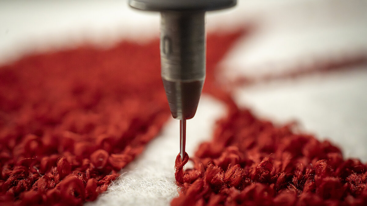 Nahaufnahme einer roten Chenille-Stickerei auf weißem Stoff, die die spezielle Chenille-Nadel zeigt, mit der der charakteristische Schlingstich gebildet wird.
